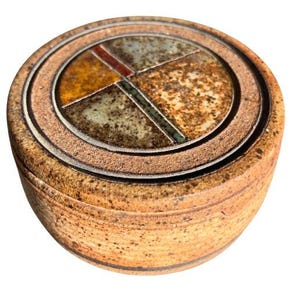 May include: A round, ceramic trinket box with a lid. The lid features a geometric design with segments of orange, brown, green, and gray. The box has a textured, speckled, brown exterior.