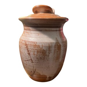 May include: A ceramic jar with a lid, featuring a rounded body and a tapered neck. The surface displays a two-tone glaze with a warm terracotta brown at the top and base, transitioning into a creamy white centre with a dark brown wheat stalk motif.