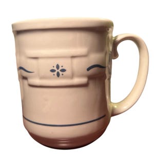 Pode incluir: Uma caneca de cerâmica na cor creme com uma faixa retangular em relevo ao redor do centro. O design inclui um motivo floral azul-escuro centralizado na faixa, ladeado por duas linhas onduladas horizontais e uma linha reta perto da base.