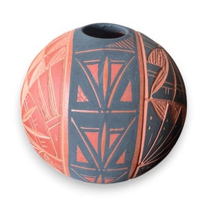 Puede incluir: Un jarrón de cerámica redondo, tallado a mano, con una combinación de colores gris oscuro y coral. El jarrón presenta patrones geométricos y florales estilizados. La parte superior tiene una pequeña abertura. Es un objeto decorativo.