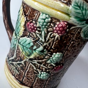 May include: A ceramic pitcher with a dark brown, textured exterior resembling tree bark. The pitcher is adorned with green leaves and clusters of red and green berries. The handle is a dark brown color.