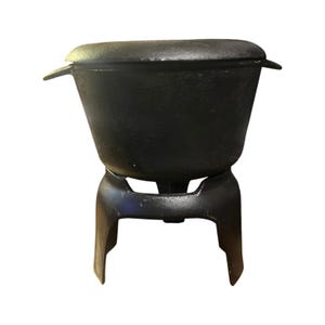 May include: A black cast iron pot with a matching lid and two side handles, resting on a sturdy three-legged cast iron stand. The pot features a wide, tapered body and a smooth, dark finish, providing a stable base for the heavy cooking vessel.