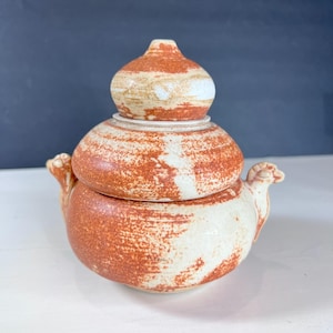 Puede incluir: Un tarro de cerámica hecho a mano con tapa. El tarro es una mezcla de blanco y marrón rojizo, con un aspecto texturizado y moteado. La tapa tiene forma bulbosa y el tarro tiene dos asas decorativas con forma de cabeza de carnero.