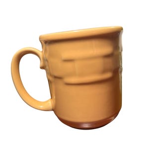 Pode incluir: Uma caneca de cerâmica de cor laranja claro sólida com um acabamento liso e brilhante e uma alça arredondada no lado esquerdo. A superfície externa da caneca apresenta um padrão horizontal em relevo sutil e repetitivo no corpo cilíndrico.