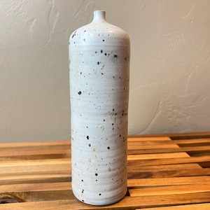 May include: A tall, cylindrical ceramic vase with a narrow neck and a rounded top. The surface is off-white with a speckled pattern featuring dark brown and tan spots. The body of the vase has subtle horizontal ridges along its entire length.