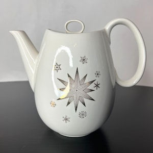 May include: Cream-coloured ceramic teapot featuring a gold and black star and snowflake pattern. The teapot has a curved spout, a large handle, and a small loop on the lid. The teapot is a winter holiday theme.