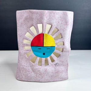 May include: A rectangular, speckled, light purple ceramic vase with a sun design. The sun has a red and yellow top half, a blue bottom half, and white rays. The vase has a unique, textured surface and a hand-painted design.