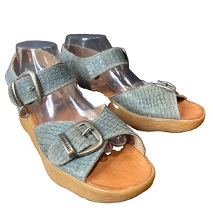 May include: A pair of light blue sandals with a textured crocodile-embossed pattern, featuring two adjustable straps with metallic buckles stamped with the word FAMOLARE, a tan leather footbed, and a thick, ridged light brown platform sole.
