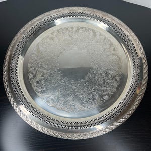 Puede incluir: Una bandeja redonda de color plateado con intrincados diseños florales y de volutas. La bandeja tiene un borde calado decorativo y un borde elevado. La bandeja es de metal y probablemente se utiliza para servir alimentos o exhibir artículos.