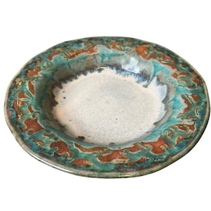 May include: A round ceramic bowl with a wide, flat rim. The center of the bowl is a light, speckled off-white color. The rim features a textured, mottled pattern of deep turquoise blue and earthy reddish-brown glaze with visible color blending.