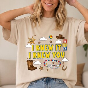 May include: A beige t-shirt featuring a graphic print with the yellow text I KNEW IT I KNEW YOU. The design includes a sheriff star, a brown cowboy hat, a pair of brown cowboy boots, a colorful beaded bracelet, alphabet blocks, and clouds.