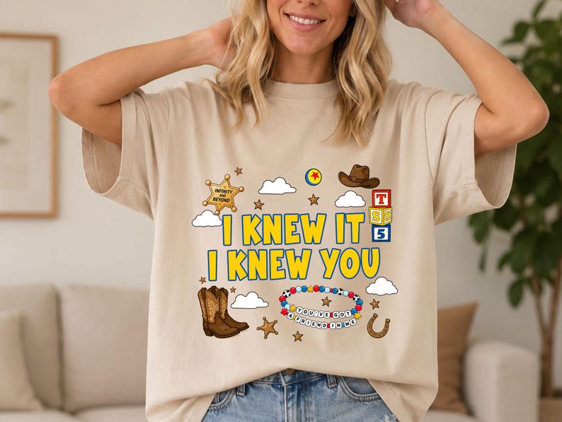 May include: A beige t-shirt featuring a graphic print with the yellow text I KNEW IT I KNEW YOU. The design includes a sheriff star, a brown cowboy hat, a pair of brown cowboy boots, a colorful beaded bracelet, alphabet blocks, and clouds.
