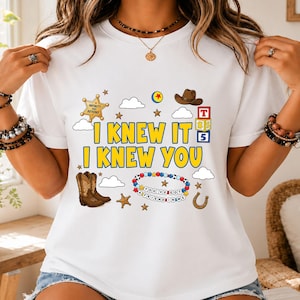 May include: A white t-shirt featuring a graphic print with the yellow text I KNEW IT I KNEW YOU. The design includes a gold sheriff badge, a brown cowboy hat, a pair of brown cowboy boots, alphabet blocks, clouds, stars, and a beaded bracelet.