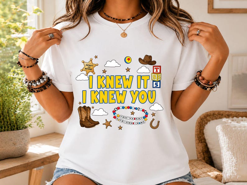 May include: A white t-shirt featuring a graphic print with the yellow text I KNEW IT I KNEW YOU. The design includes a gold sheriff badge, a brown cowboy hat, a pair of brown cowboy boots, alphabet blocks, clouds, stars, and a beaded bracelet.