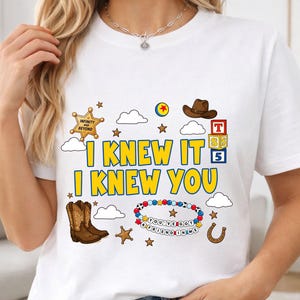 May include: A white t-shirt featuring a graphic print with the yellow text I KNEW IT I KNEW YOU. The design includes a brown cowboy hat, brown cowboy boots, a sheriff badge, a colorful beaded bracelet with the text YOU'VE GOT A FRIEND IN ME, and blocks.
