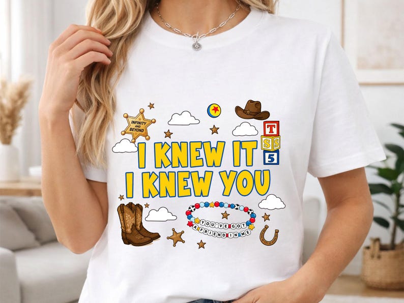 May include: A white t-shirt featuring a graphic print with the yellow text I KNEW IT I KNEW YOU. The design includes a brown cowboy hat, brown cowboy boots, a sheriff badge, a colorful beaded bracelet with the text YOU'VE GOT A FRIEND IN ME, and blocks.