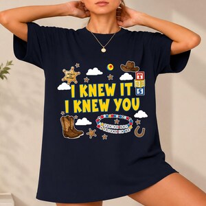 May include: A dark navy blue t-shirt featuring the yellow text I KNEW IT I KNEW YOU. The graphic includes a sheriff badge, a brown cowboy hat, alphabet blocks, a beaded bracelet, a horseshoe, stars, and clouds in a colorful, playful design.