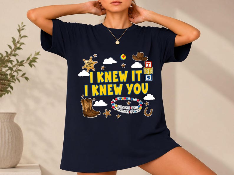 May include: A dark navy blue t-shirt featuring the yellow text I KNEW IT I KNEW YOU. The graphic includes a sheriff badge, a brown cowboy hat, alphabet blocks, a beaded bracelet, a horseshoe, stars, and clouds in a colorful, playful design.