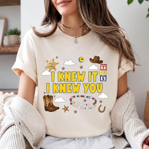 May include: A cream-colored t-shirt featuring a graphic print with the text I KNEW IT I KNEW YOU. The design includes a sheriff badge, a brown cowboy hat, building blocks, cowboy boots, a beaded bracelet with the text YOU'VE GOT A FRIEND IN ME.