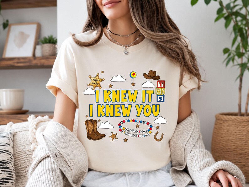 May include: A cream-colored t-shirt featuring a graphic print with the text I KNEW IT I KNEW YOU. The design includes a sheriff badge, a brown cowboy hat, building blocks, cowboy boots, a beaded bracelet with the text YOU'VE GOT A FRIEND IN ME.