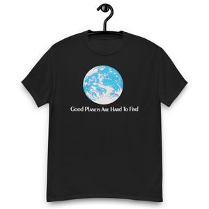 May include: A black short-sleeve t-shirt featuring a circular graphic of the Earth in shades of blue and white. Below the planet, the text "Good Planets Are Hard To Find" is printed in a white serif font. The shirt is displayed on a black hanger.