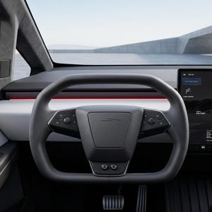 May include: The interior of a vehicle featuring a dark gray yoke-style steering wheel with integrated controls, a minimalist dashboard with a horizontal red light strip, and a large touchscreen display showing a vehicle graphic and various settings.