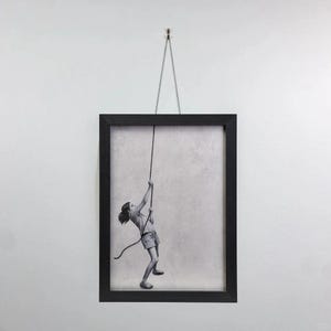 May include: Black framed art print of a child climbing a rope. The black and white image shows a young person in shorts and a t-shirt, grasping a rope and ascending. The artwork is suspended by a thin wire from a wall.