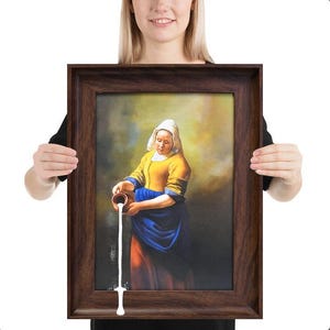 Könnte beinhalten: Gerahmter Kunstdruck eines Gemäldes, das eine Frau zeigt, die Milch aus einem Krug gießt. Die Frau trägt ein gelbes Oberteil, einen blauen Rock und eine weiße Kopfbedeckung. Der Rahmen ist dunkelbraun.