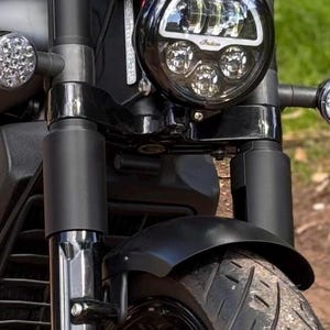 May include: A close-up shot of a black motorcycle's front end. The image focuses on the headlight, front tyre, mudguard, and suspension. The round headlight has multiple LED lights. The tyre is black with a tread pattern. The mudguard is curved and black.