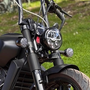 May include: A close-up shot of a black motorcycle's front end. It has a round LED headlight, chrome handlebars, and a black front mudguard. The tyre is black with a tread pattern, set against a backdrop of green grass.