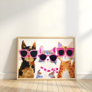 Könnte beinhalten: Ein gerahmter Kunstdruck mit drei stilisierten Katzen, die rosa Sonnenbrillen tragen. Die Katzen haben unterschiedliche Fellmuster in Braun-, Blau- und Orangetönen. Das Kunstwerk ist vor einem weißen Hintergrund in hellem Holz gerahmt.