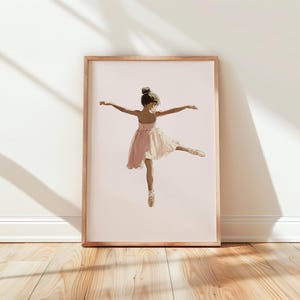 May include: A framed print of a young ballerina in a pink dress and ballet shoes. The artwork depicts the dancer mid-leap with arms outstretched. The background is a soft pink, and the frame is a light wood color.