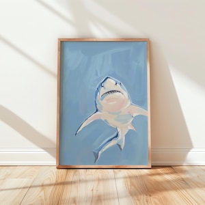 May include: A framed art print featuring a white and blue shark swimming in a light blue background. The shark is painted with soft brushstrokes, and the frame is a natural wood color. The artwork is displayed against a white wall and wooden floor.