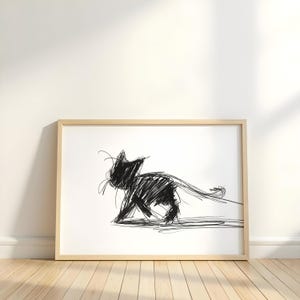 Könnte beinhalten: Ein gerahmter Kunstdruck mit einer schwarzen Katzenzeichnung auf weißem Hintergrund. Die Katze ist in einem minimalistischen Stil mit kräftigen, gekritzelten Linien dargestellt. Der Rahmen ist helles Holz, und das Kunstwerk wird auf einem Holzboden präsentiert.