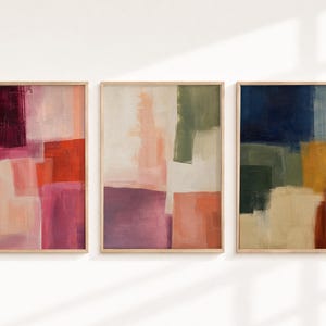 Pode incluir: Um conjunto de três impressões de arte de parede retangulares em molduras de madeira clara. A primeira impressão apresenta blocos de bordô profundo, rosa e laranja. A segunda exibe creme, pêssego e verde-oliva. A terceira mostra azul-marinho, mostarda e castanho.