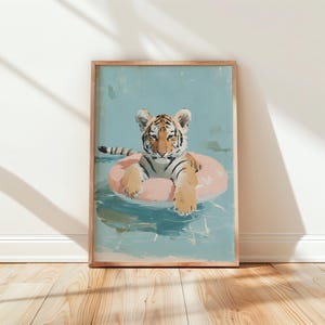 Op de afbeelding: Een ingelijste kunstdruk met een tijgerwelp die in een roze opblaasbare ring in een zwembad drijft. Het kunstwerk heeft een lichtblauwe achtergrond met waterreflecties. De tijger heeft oranje en zwarte strepen.