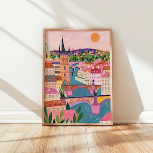 Op de afbeelding: Een ingelijste kunstdruk met een kleurrijk stadsgezicht met een rivier, bruggen en gebouwen. Het kunstwerk heeft een warm kleurenpalet met roze, oranje en blauwe tinten. Een heldere oranje zon staat aan de hemel.