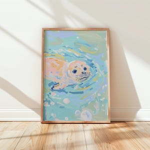 Puede incluir: Una impresión artística enmarcada que representa una foca nadando en el agua. La foca está pintada en tonos melocotón y crema, con ojos azules y bigotes. El agua está representada en tonos azules y verdes, con burbujas blancas. El marco es de color marrón claro.