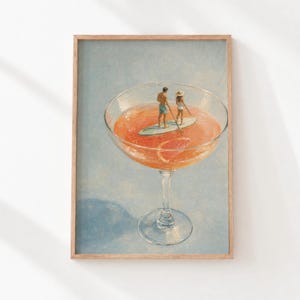 May include: A framed art print featuring a large cocktail glass filled with orange liquid and a grapefruit slice. Inside the glass, two small figures stand on a paddleboard, with one person holding a paddle, set against a light blue background.