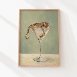以下が含まれることがあります： 大きなマティーニグラスの中で休むヒョウの絵が描かれた額装アートプリント。ヒョウは茶色とベージュの斑点模様の毛皮をしており、グラスの底には爪楊枝に刺さった緑色のオリーブが一つ入っています。背景は無地の落ち着いたセージグリーン色です。