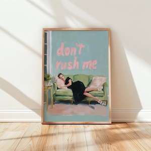 May include: A framed art print depicting a woman lounging on a green sofa. The artwork has a light blue backdrop with the words "don't rush me" in pink. The woman wears a black dress and white shoes.