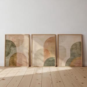 May include: Three framed abstract art prints. Each print features a beige background with overlapping semi-circular shapes in shades of brown, green, and peach. The frames are a light wood colour, and the artwork is displayed on a light-coloured wall.