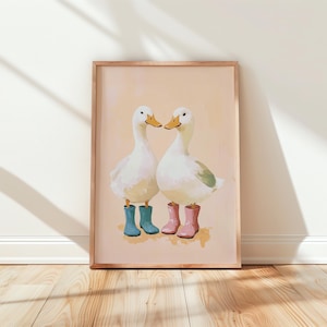 May include: A framed print featuring two white ducks with yellow beaks and wearing blue and pink rain boots. The ducks are facing each other on a light peach background. The artwork is in a wooden frame.