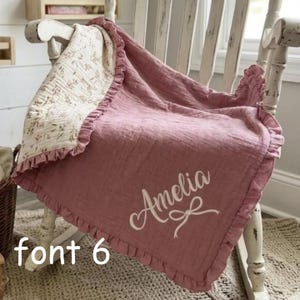 May include: A dusty rose muslin blanket with a ruffled edge and a cream-coloured floral patterned reverse side. The blanket features the name Amelia in white cursive script with a small bow graphic. The text font 6 is visible in the corner.