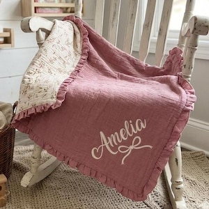 May include: A dusty rose-colored blanket with a ruffled edge and the name Amelia embroidered in white script with a small bow detail. The reverse side of the blanket features a cream-colored fabric with a delicate brown floral pattern.
