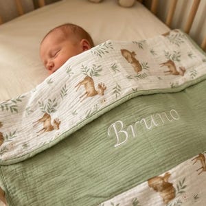 May include: A soft muslin baby blanket featuring a white background with a repeating pattern of brown deer and green foliage, accented by a solid sage green section embroidered with the name Bruno in white cursive script lettering.
