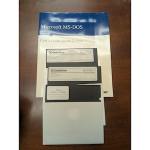 Op de afbeelding: Een Microsoft MS-DOS gebruikershandleiding en naslagwerk vergezeld van drie zwarte 3,5-inch diskettes. De diskettes zijn gelabeld met GoldStar MS-DOS 5, inclusief Disk 1 van 2, Disk 2 van 2 en een Mouse Driver & Utilities disk in witte hoesjes.