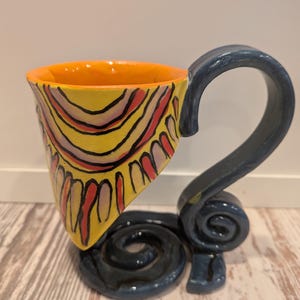 May include: A unique ceramic mug with a yellow, orange, and blue color scheme. The mug features a distinctive handle and base design. The cup's exterior is decorated with a yellow background and red and black curved lines.
