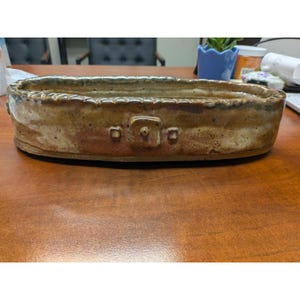 May include: An elongated oval ceramic tray with a mottled tan, brown, and cream glaze. The rim features a scalloped texture, and the exterior side displays a central square relief detail with a small raised dot flanked by two smaller squares.