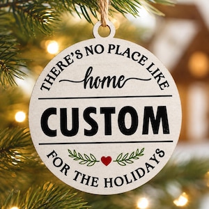 Op de afbeelding: Een rond, lichtgekleurd houten ornament dat aan een touwtje van jute hangt. De tekst luidt "THERE'S NO PLACE LIKE home" in het zwart, gevolgd door "CUSTOM" in grote, dikgedrukte letters en "FOR THE HOLIDAYS" met een klein rood hartje en groene blaadjes.
